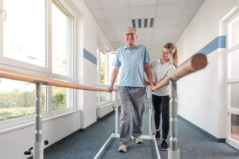 Smiling elderly man practicing walking between parallel bars with the assistance of a physiotherapist in a bright rehabilitation facility.