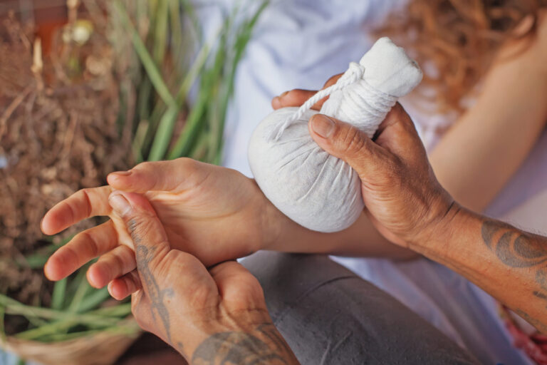 Close-up of a therapist applying a herbal clay hot pack to a person’s wrist during a massage therapy session, focusing on relaxation and localized pain relief.