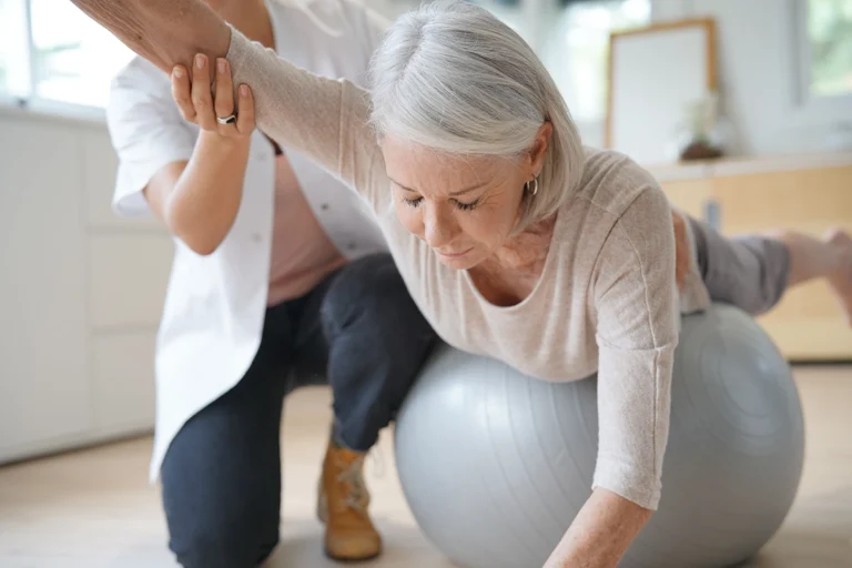 Senior woman balancing on a stability ball while reaching forward with assistance from a physiotherapist, practicing strength and coordination exercises in a bright therapy room.