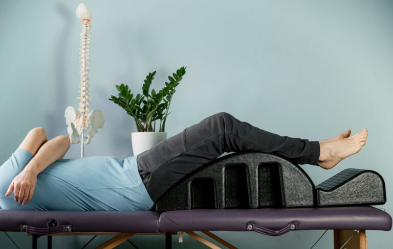 Person lying on a treatment table with legs elevated on a lumbar support wedge, used for spine decompression or therapeutic back positioning, with an anatomical spine model and plant in the background.