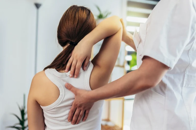 Physiotherapist guiding a woman through a shoulder mobility exercise, with one hand supporting her back and the other adjusting her arm in a clinical environment.