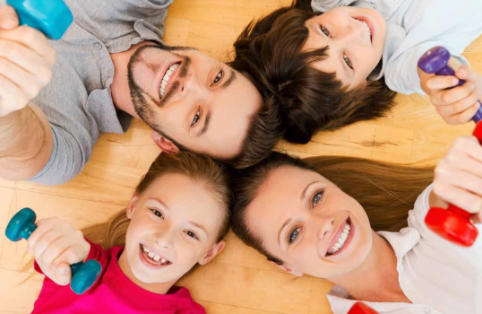 Happy family of four lying on the floor in a circle, smiling up at the camera while holding colorful hand weights, promoting fun and fitness together.