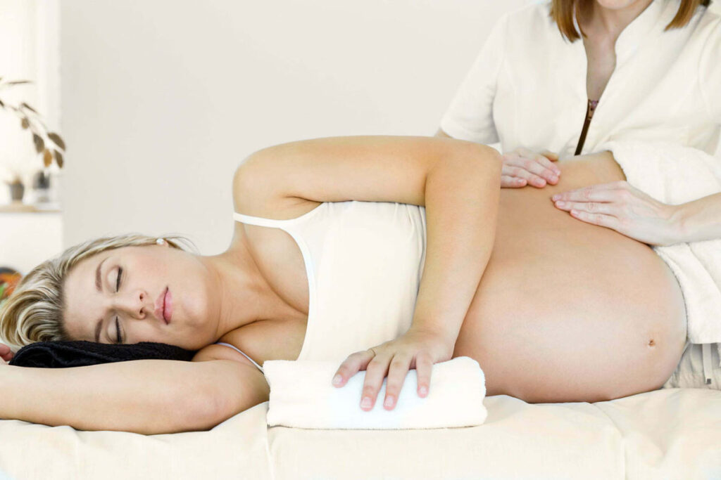 Pregnant woman lying on her side with eyes closed, receiving a gentle abdominal massage from a massage therapist in a calm, supportive environment.